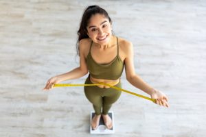 Indian female measuring waist while standing on scales, happy about successful weight loss result at